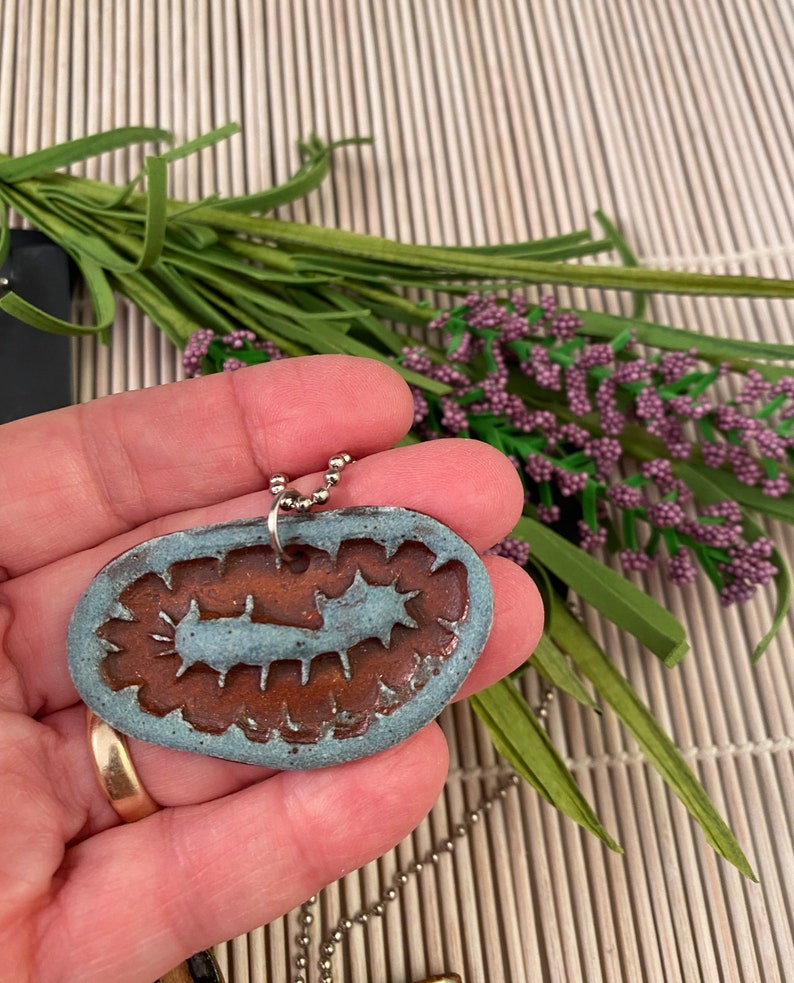 May include: A blue and brown ceramic pendant with a detailed design, hanging from a silver chain. The pendant is shaped like a worm or caterpillar.