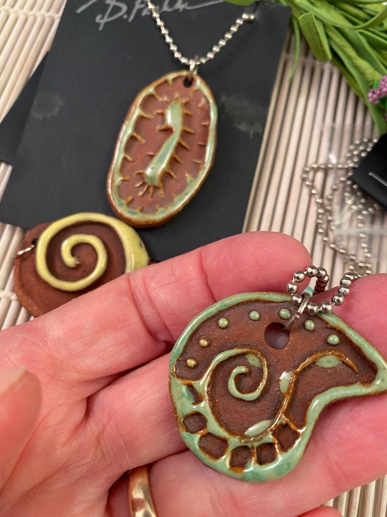 May include: Three ceramic pendants with brown and green glaze. The pendants feature different designs, including a swirl, a leaf, and a geometric pattern. The pendants are on silver chains.