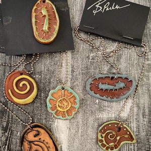 May include: Five ceramic pendants on silver chains. The pendants are all different shapes and sizes, and feature various designs in brown, green, and yellow. The pendants are all signed "B. Parker".