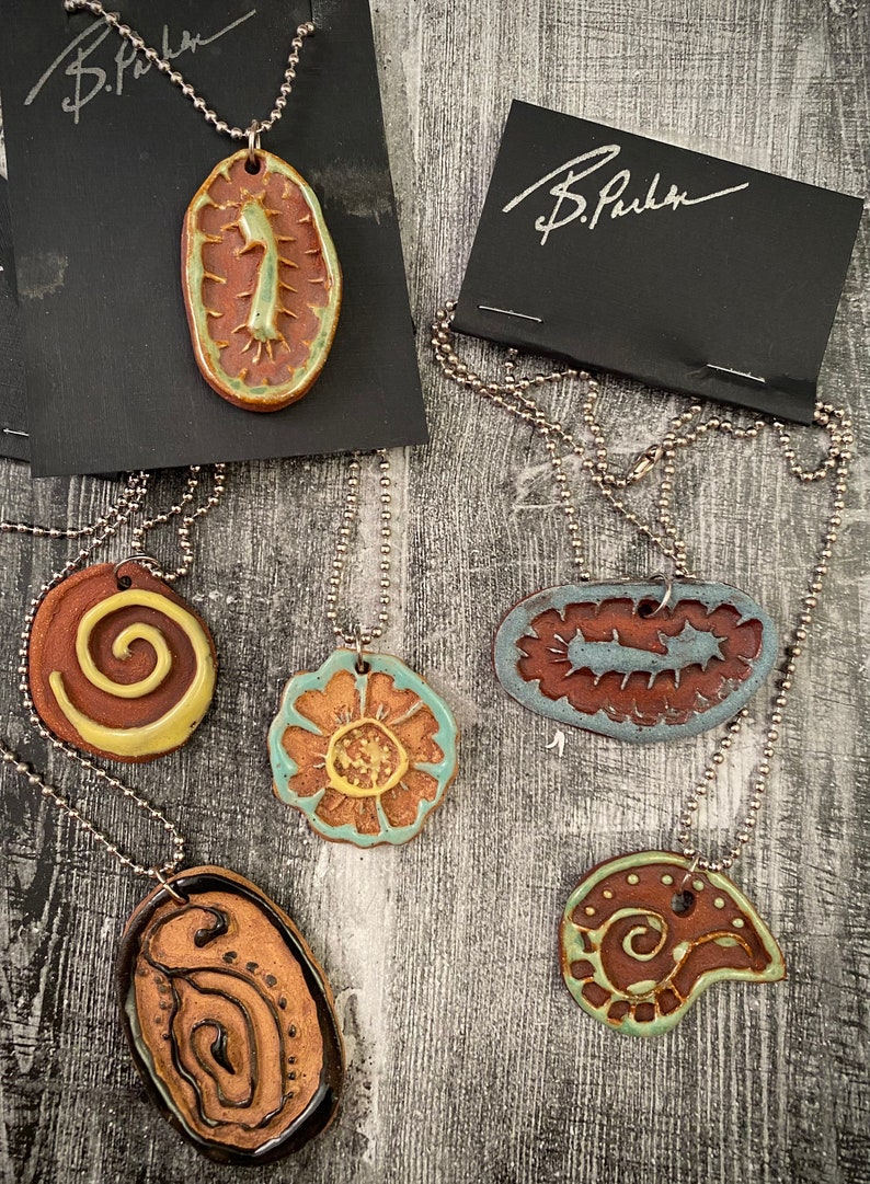 May include: Five ceramic pendants on silver chains. The pendants are all different shapes and sizes, and feature various designs in brown, green, and yellow. The pendants are all signed "B. Parker".