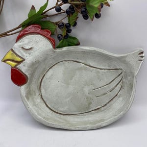 May include: A ceramic hen-shaped serving dish. The dish is primarily gray with a red comb and wattle, and a yellow beak. The dish has a brown rim and etched wing details. Decorative berries and leaves are in the background.