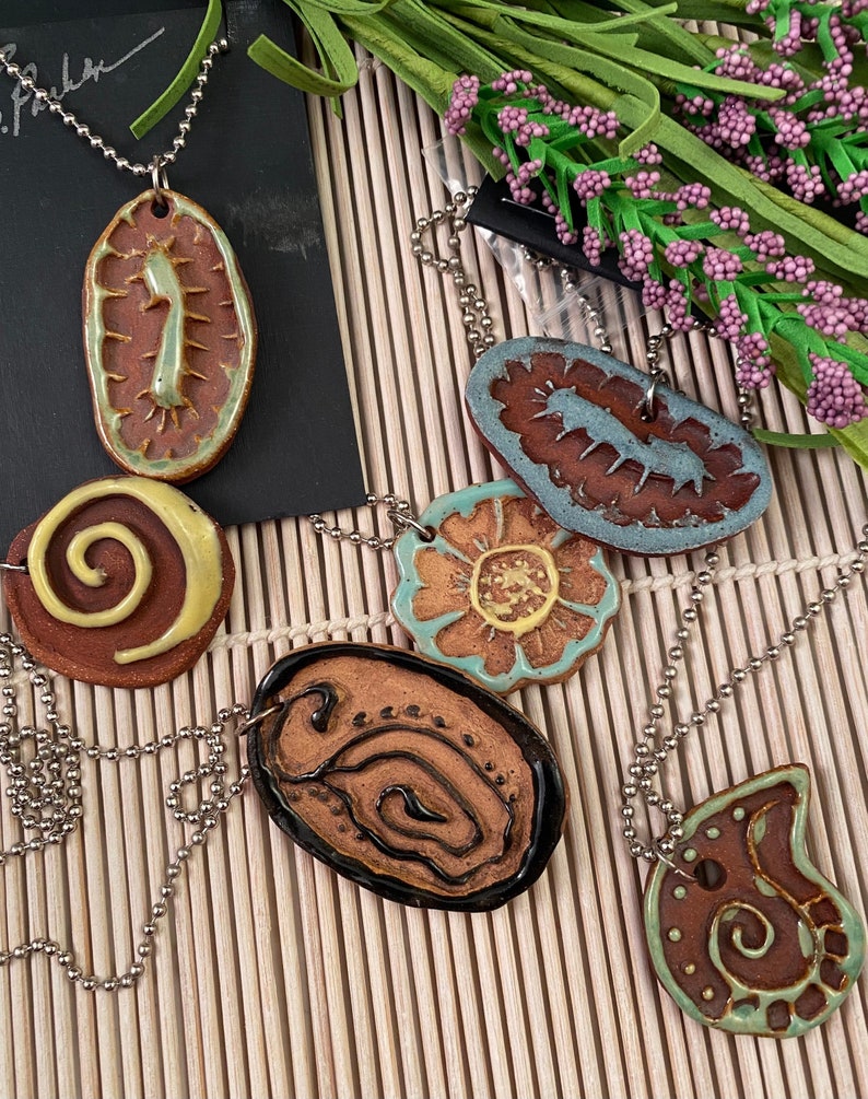May include: Five ceramic pendants with different designs, including a spiral, a flower, and a paisley pattern. The pendants are strung on silver chains.