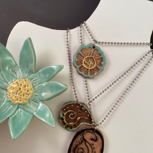 May include: Three ceramic pendants on silver chains. The pendants are a brown and turquoise paisley design, a brown and turquoise flower, and a brown and black oval with a swirl design.