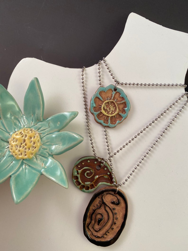 May include: Three ceramic pendants on silver chains. The pendants are a brown and turquoise paisley design, a brown and turquoise flower, and a brown and black oval with a swirl design.