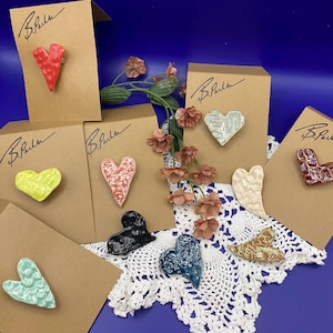 May include: Assortment of handmade ceramic heart-shaped pins in various colors and patterns, including red, green, blue, pink, and white. The pins are displayed on brown cards with the text "B.Puhn" written on them.