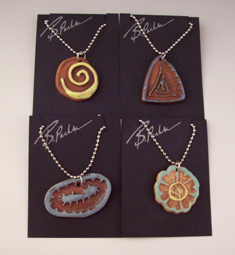 May include: Four ceramic pendants on silver chains, each with a different design: a spiral, a triangle, a flower, and a wavy line. The pendants are displayed on black cards with the signature "B. Parker" on each.