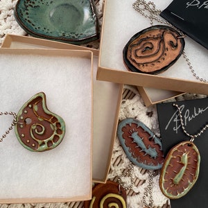May include: A set of five ceramic pendants with different designs, including a heart, a swirl, a flower, and two abstract shapes. The pendants are on chains and are displayed in boxes with a signature.