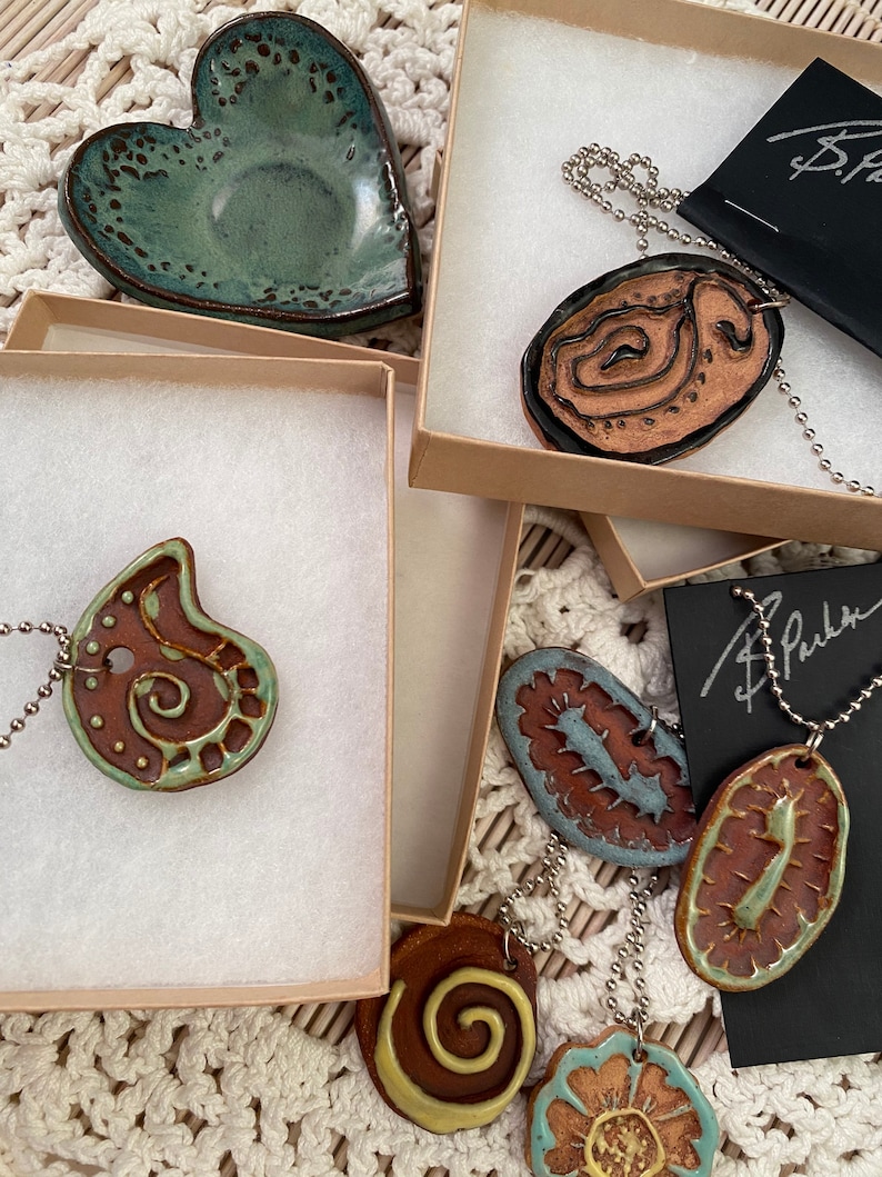 May include: A set of five ceramic pendants with different designs, including a heart, a swirl, a flower, and two abstract shapes. The pendants are on chains and are displayed in boxes with a signature.
