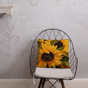 May include: A throw pillow with a close-up image of three sunflowers with yellow petals and dark brown centers. The pillow is sitting on a white chair with a black metal frame.