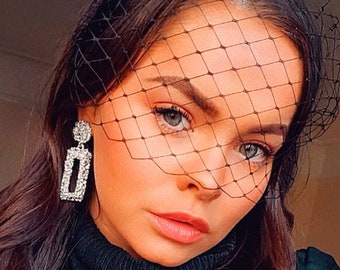 May include: A woman with long dark hair wearing a black veil over her face. She is wearing a dark green turtleneck sweater and has large rectangular silver earrings with rhinestones.