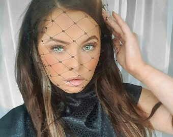 May include: A woman with long brown hair wearing a black mock neck shirt with a subtle snake skin pattern. She is wearing a black veil with a diamond pattern.