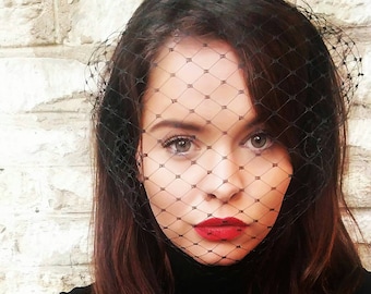 May include: A woman with long dark brown hair is wearing a black veil over her face. She is wearing a black turtleneck sweater and has red lipstick on.