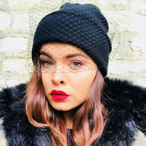 以下が含まれることがあります： 顔に黒いメッシュベールをつけた黒いビーニーをかぶった女性。彼女は黒いファーのトリミングが施されたジャケットを着ており、赤い口紅をしています。