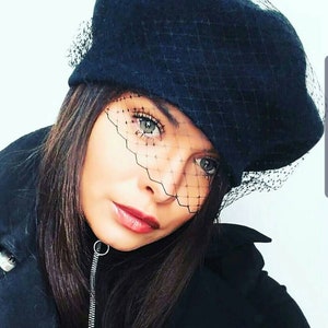 May include: A woman wearing a black beret with a black mesh veil. The veil covers her eyes and part of her face. She is wearing a black jacket.