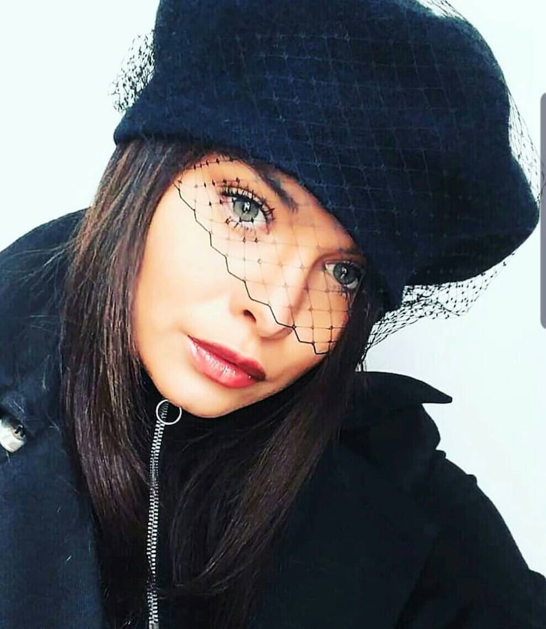 May include: A woman wearing a black beret with a black mesh veil. The veil covers her eyes and part of her face. She is wearing a black jacket.