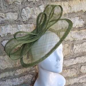 Op de afbeelding: Een groene, breedgerande hoed met een grote strik aan de zijkant. De hoed is gemaakt van een delicate, gaasachtige stof.