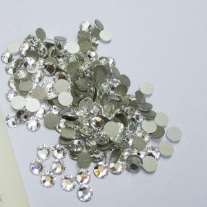 May include: A pile of clear glass rhinestones with silver adhesive backs. The rhinestones are scattered on a white surface.