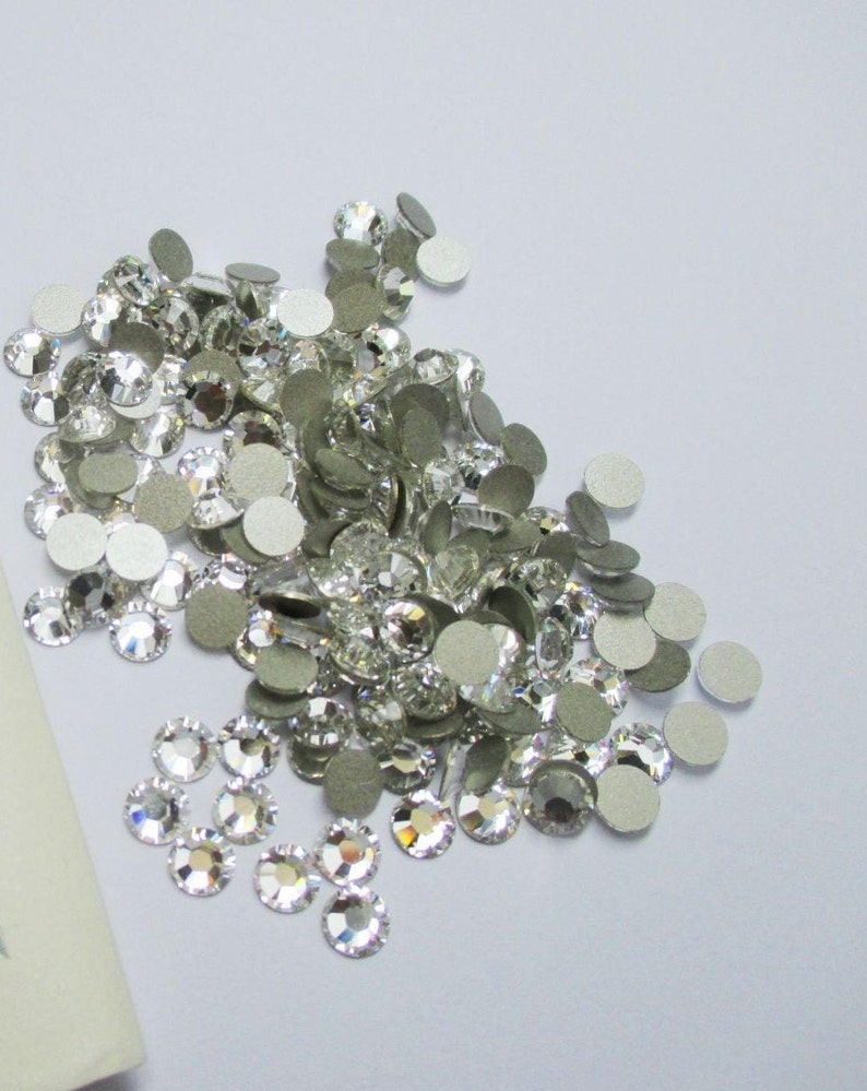 May include: A pile of clear glass rhinestones with silver adhesive backs. The rhinestones are scattered on a white surface.