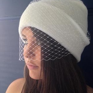 girl wearing pure white ribbed smooth beanie hat, folded cuff style and delicate vintage style fascinator veil to front to mask eyes