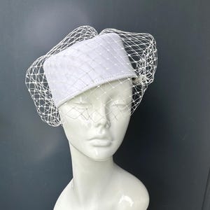ベール付きの白いピルボックス型スチュワーデス風カクテルハット。結婚式やフォーマルな場面にふさわしい帽子です。