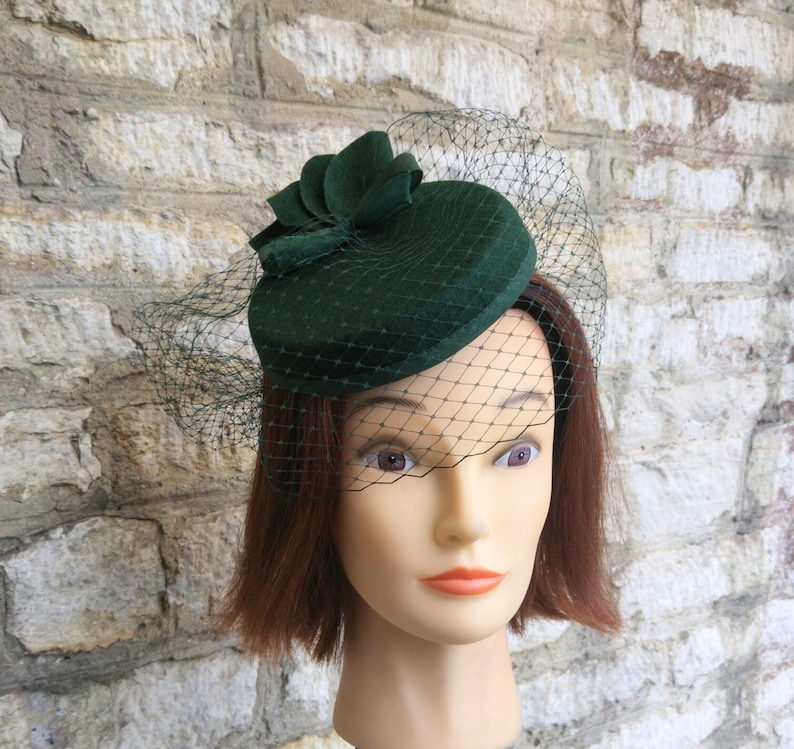 May include: A dark green hat with a flower accent and a delicate net veil. The hat is a deep forest green color and sits atop a mannequin head. The veil is a fine mesh, adding a touch of elegance.