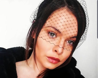 May include: A woman wearing a black veil over her face. The veil is made of a fine mesh material and covers her forehead, eyes, and nose. Her hair is pulled back and she is wearing a black jacket.