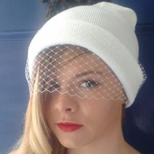 blonde girl red lipstick in white beanie hat with delicate fascinator veil to front, shielding eyes