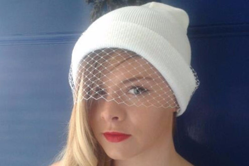 blonde girl red lipstick in white beanie hat with delicate fascinator veil to front, shielding eyes