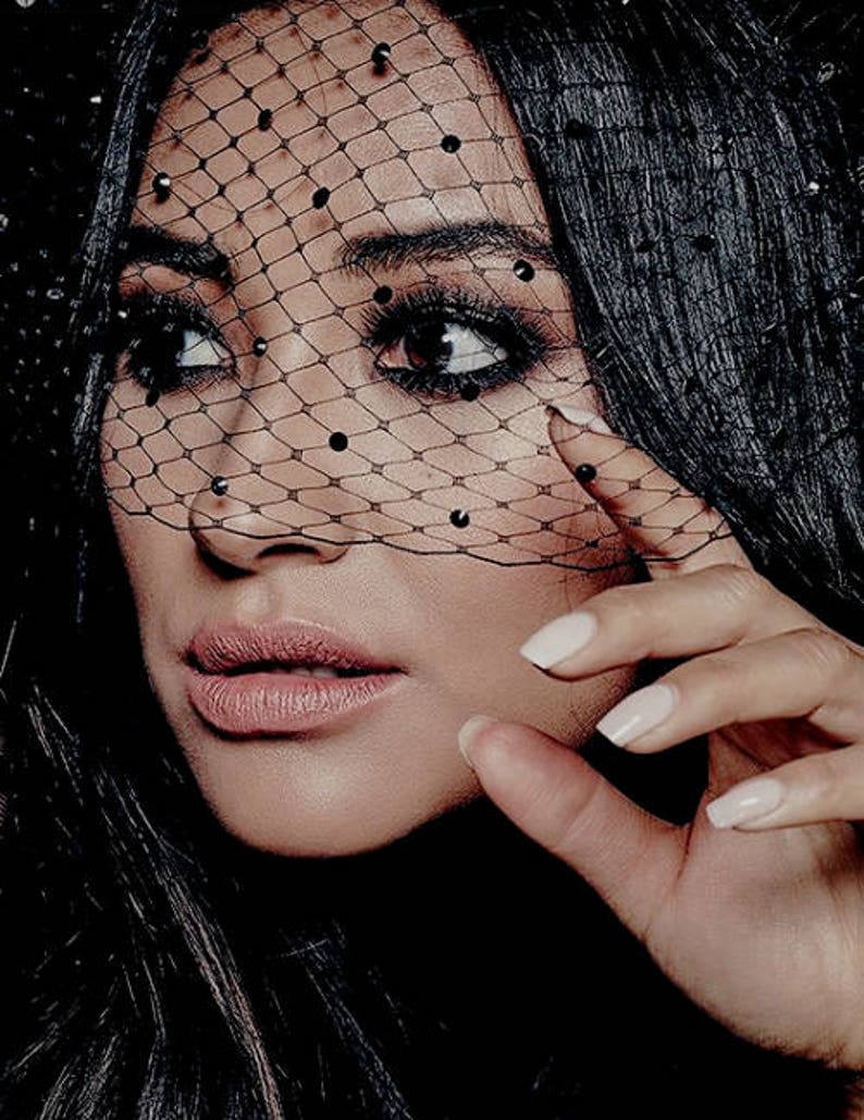 May include: A woman with long black hair wearing black eye makeup and a black veil with small black dots. Her nails are painted white.