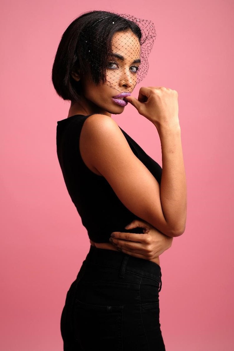 May include: A woman with short black hair is wearing a black sleeveless top and black trousers. She is wearing a black veil with rhinestones over her face. She is looking at the camera with a serious expression.