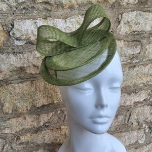 May include: A green, layered, circular hat with a large bow on top. The hat is made of a woven material and has a delicate, airy feel.