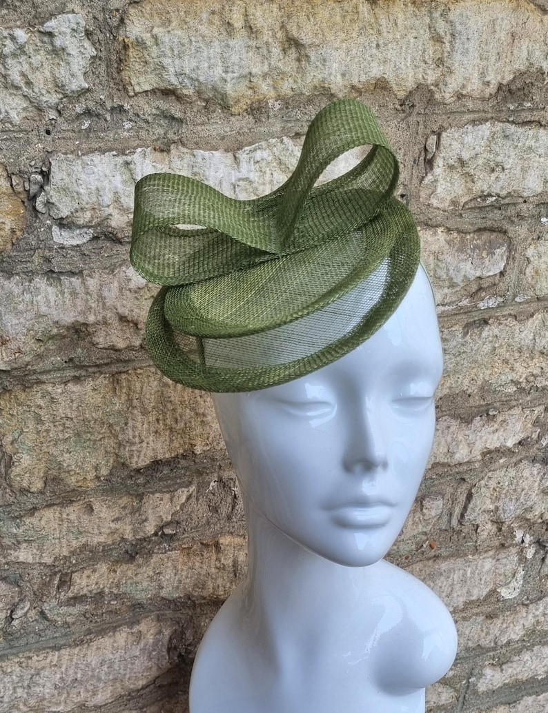 May include: A green, layered, circular hat with a large bow on top. The hat is made of a woven material and has a delicate, airy feel.