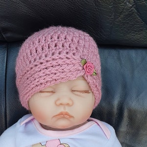 May include: A pink crocheted baby hat with a small pink flower embellishment. The hat is angled on the head of a sleeping baby doll.