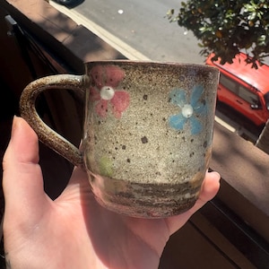 Puede incluir: Taza de cerámica hecha a mano con un tono terroso moteado. La taza presenta diseños florales pintados a mano en rosa y azul, con centros blancos. El asa y el borde tienen un acabado más oscuro y texturizado, lo que añade encanto rústico.