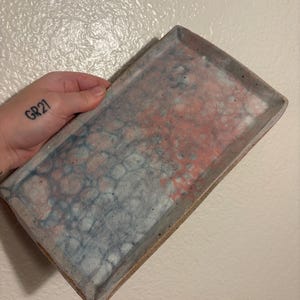 Op de afbeelding: Rechthoekige keramische schaal met een gespikkeld ontwerp in tinten rood, blauw en grijs. De schaal heeft een opstaande rand en een natuurlijke, ongeglazuurde rand. Het interieur heeft een uniek, abstract patroon.