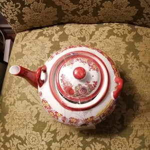May include: A white teapot with a red lid and floral designs. The teapot has a red handle and spout. The teapot is sitting on a green and gold patterned surface.