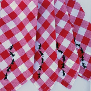 May include: Four red and white gingham napkins with black embroidered ants. The napkins are folded in half and arranged in a fan shape.
