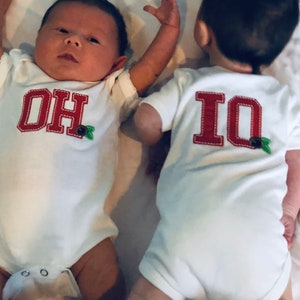 Puede incluir: Dos bebés con monos blancos con letras bordadas en rojo. Un mono dice "OH" y el otro dice "IO".