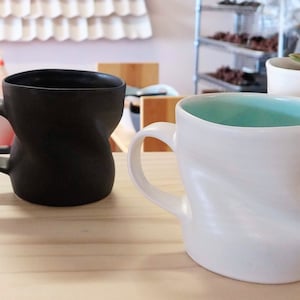 May include: Two ceramic mugs with a unique, wavy design. One mug is black and the other is white with a light blue interior.