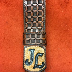 May include: A brown leather belt with a geometric pattern and the initials "JL" in a gold and black design. The belt is made of a dark brown leather with a repeating square pattern.