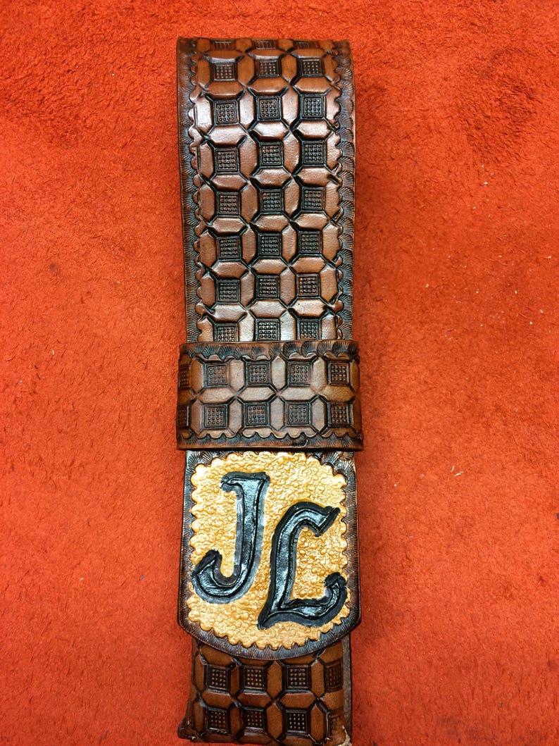 May include: A brown leather belt with a geometric pattern and the initials "JL" in a gold and black design. The belt is made of a dark brown leather with a repeating square pattern.