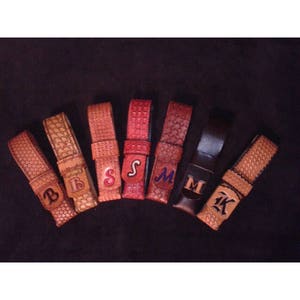 May include: A collection of leather belt keepers in various shades of brown and red, each embossed with a different letter. The letters are in different fonts and colors. The keepers are arranged on a dark background.