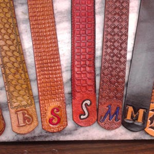 May include: A collection of leather belts in various shades of brown, red, and black, each with a unique embossed pattern. Several belts feature initials such as "B", "S", "M", and "K" in contrasting colors, showcasing personalized craftsmanship.