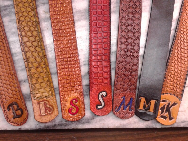 May include: A collection of leather belts in various shades of brown, red, and black, each with a unique embossed pattern. Several belts feature initials such as "B", "S", "M", and "K" in contrasting colors, showcasing personalized craftsmanship.