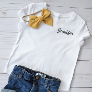 May include: White t-shirt with the name "Jennifer" embroidered on the front. The shirt is paired with denim shorts and a yellow polka dot headband.