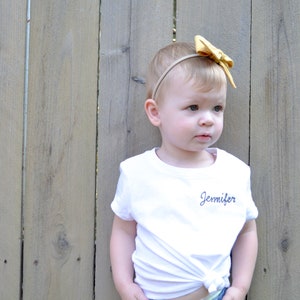 May include: A young child wearing a white t-shirt with the name "Jennifer" embroidered on the front. The child is wearing a yellow bow headband and blue jeans. The child is standing in front of a wooden fence.