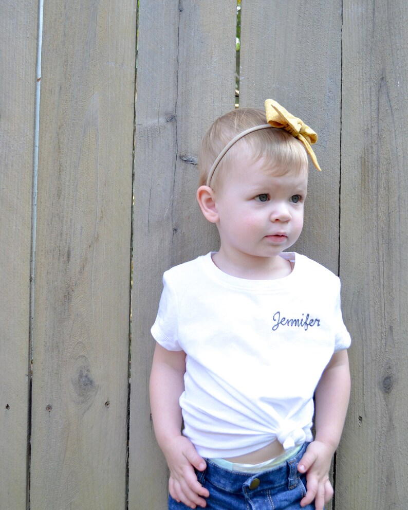 May include: A young child wearing a white t-shirt with the name "Jennifer" embroidered on the front. The child is wearing a yellow bow headband and blue jeans. The child is standing in front of a wooden fence.