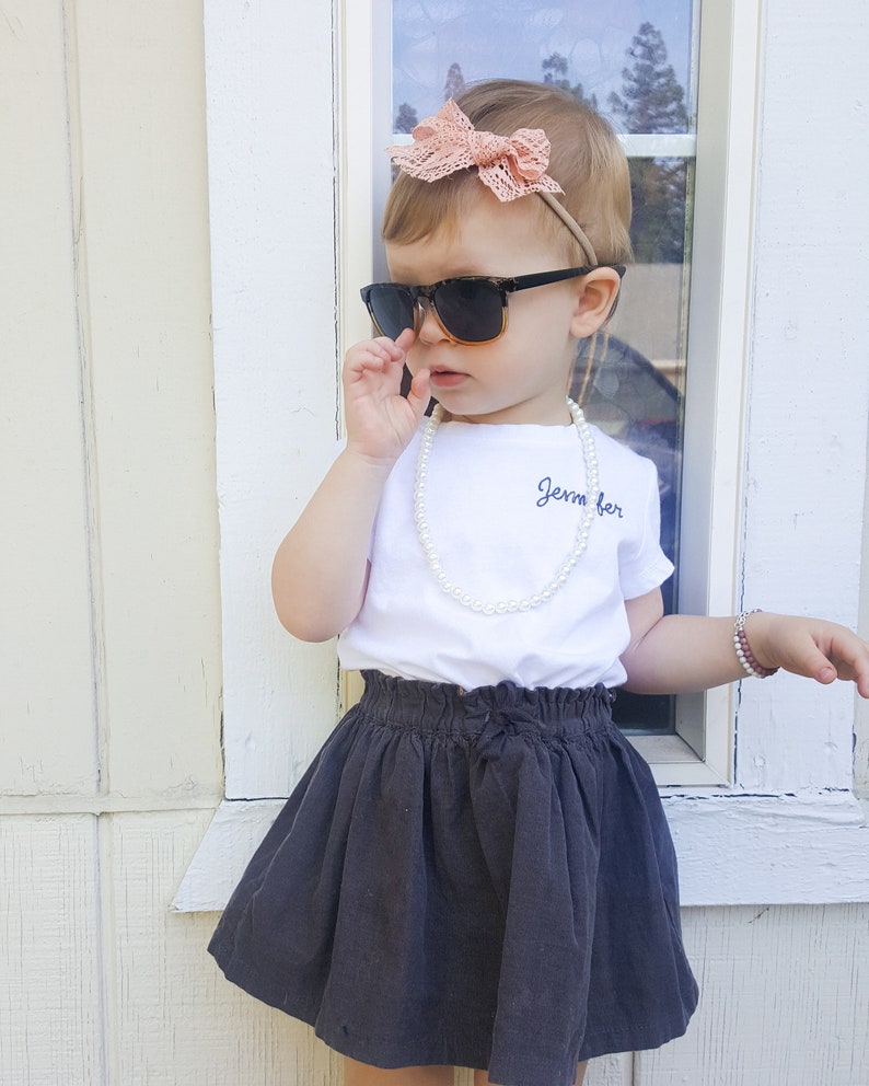 May include: A young girl wearing a white t-shirt with the text "Jemma" embroidered on it, a black skirt, a pearl necklace, sunglasses, and a pink bow headband.
