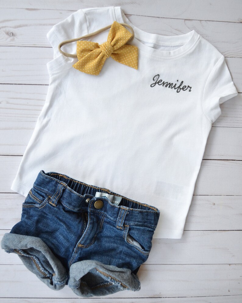 May include: White t-shirt with the name "Jennifer" embroidered on the front. The shirt is paired with denim shorts and a yellow polka dot headband.