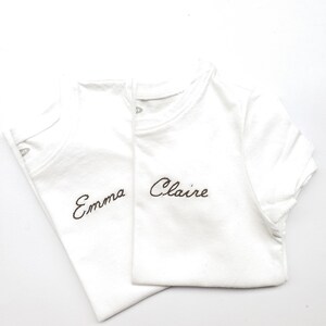 May include: Two white t-shirts with brown embroidered names. The top shirt says "Claire" and the bottom shirt says "Emma".